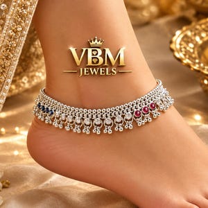 May include: A silver anklet featuring a detailed beaded chain with small dangling charms, accented by alternating blue and red circular stones. The anklet is marked with the text VBM JEWELS and a crown icon, showcasing a metallic finish.