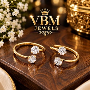 Op de afbeelding: Twee open gouden ringen, elk voorzien van twee ronde briljant geslepen heldere stenen aan de uiteinden van de band. De tekst VBM JEWELS met een kroonpictogram staat erboven. De sieraden liggen op een donker gepolijst houten oppervlak.