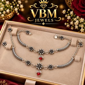 May include: A pair of silver-toned anklets featuring intricate floral motifs, textured cylindrical links, and small dangling red beads. The jewellery is displayed in a rectangular box with the text VBM JEWELS and a crown logo visible above.