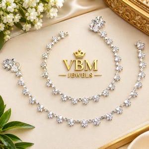 May include: Two silver-toned anklets featuring a repeating floral pattern set with clear, sparkling stones. Each anklet has a larger floral clasp at one end. The text VBM JEWELS with a crown icon is centred between the two pieces of jewellery.