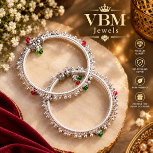 May include: Two circular silver bangles featuring intricate metalwork and small dangling beads in red and green. The jewelry is branded with the text VBM Jewels and includes icons for premium quality, 925 sterling silver, and skin friendly.