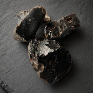 Op de afbeelding: Drie ruwe, ongepolijste obsidiaanstenen met een glanzend, gitzwart uiterlijk. Elke steen heeft onregelmatige, scherpe randen en een gladde, glasachtige oppervlaktestructuur met subtiele variaties in lichtreflectie op het donkere gesteente.