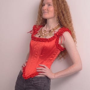 May include: A bright red satin corset top featuring vertical boning channels, a square neckline, and ruffled trim along the straps and hem. The garment includes thin ribbon ties at the shoulders and a structured, fitted bodice design.