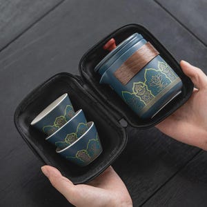 May include: A portable tea set in a black zippered carrying case, featuring a blue ceramic teapot with a wooden handle and a red lid knob, alongside three matching blue ceramic tea cups. Each piece is decorated with a gold lotus pattern.