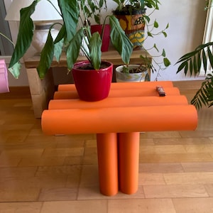 Könnte beinhalten: Ein leuchtend orangefarbener Beistelltisch, der aus mehreren zylindrischen Rohren besteht, die horizontal zur Tischplatte und vertikal zu zwei Stützbeinen angeordnet sind. Der Tisch hat eine glatte, matte Oberfläche und eine durchgehend einheitliche Farbe.