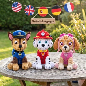 Op de afbeelding: Drie gehaakte knuffels die tekenfilmhonden voorstellen. De linkerfiguur is bruin en blauw met een politiepet. De middelste figuur is wit met zwarte vlekken en een rode brandweerhelm. De rechterfiguur is beige en roze met een roze vliegersbril.