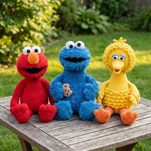 Könnte beinhalten: Drei handgefertigte Häkel-Plüschtiere, bestehend aus einer roten Figur mit großer orangefarbener Nase, einer blauen Figur, die einen kleinen braunen Keks hält, und einer gelben vogelartigen Figur mit orangefarbenen Füßen und gestreiften Beinen.