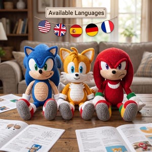 May include: Three crocheted plush toys representing Sonic, Tails, and Knuckles. The blue Sonic, yellow Tails, and red Knuckles figures are seated side-by-side. Above them, a sign reads Available Languages with five circular national flags.