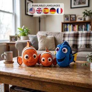 May include: Three crocheted plush fish toys sitting on a wooden table. Two are orange and white clownfish of different sizes, and one is a blue and black tang fish with large eyes. Above them, a sign reads AVAILABLE LANGUAGES with five flags.