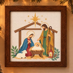 May include: A framed cross-stitch artwork depicting a nativity scene with a central star, a manger containing an infant, two figures, and a lamb. The figures are dressed in blue, red, green, and tan robes, set within a wooden structure.