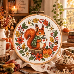 May include: A circular wooden embroidery hoop containing a cross-stitch pattern of a squirrel wearing a green scarf and holding a white mug with a red heart. The squirrel sits on a tree stump surrounded by a wreath of autumn leaves and flowers.