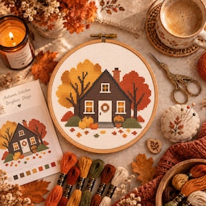 May include: A circular wooden embroidery hoop containing a cross-stitch pattern of a dark brown house surrounded by yellow and orange autumn trees. A printed pattern guide titled Autumn Stitches Brighter Days, embroidery floss, and scissors.