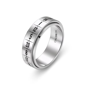 Puede incluir: Un anillo de metal plateado con una banda exterior giratoria grabada con números negros y líneas verticales. Los números 16, 12, 5, 20, 8, 17 y 2 son visibles. La palabra NUMENO está grabada en la superficie interior de la banda del anillo.