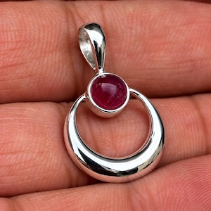 Puede incluir: Un colgante de plata con un marco metálico circular pulido de superficie lisa y reflectante. En la parte superior del círculo, una piedra preciosa cabujón redonda de color rosa intenso está engastada en un bisel de plata con una reasa.