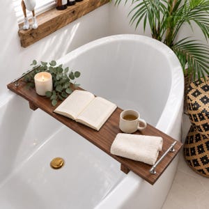 Puede incluir: Una bandeja rectangular de madera oscura para bañera con asas de metal plateado sobre una bañera blanca. La bandeja sostiene una vela blanca encendida, una rama de eucalipto verde, un libro abierto, una taza de cerámica beige y una toalla beige.