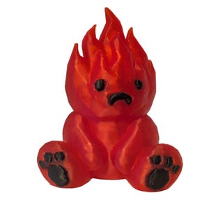 May include: A small, red, 3D-printed figurine shaped like a seated bear with a flame-like head. The figure features a sad facial expression with black eyes and a downturned mouth, along with black paw pads on the feet, all rendered in a glossy red material.