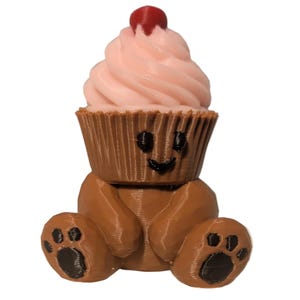 May include: A brown 3D printed bear figurine with black paw pads, featuring a cupcake head with light pink frosting and a small red cherry on top. The cupcake portion has a smiling face with black eyes and a mouth, creating a whimsical character.
