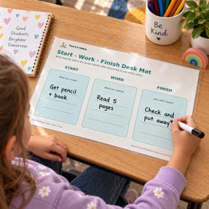 May include: A laminated Start-Work-Finish desk mat with three light blue sections labeled Start, Work, and Finish. The mat includes text prompts for tasks and is being written on with a black dry-erase marker. A notebook with heart patterns and a pencil cup are nearby.
