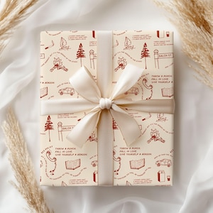 May include: A rectangular gift box wrapped in cream-colored paper with a repeating pattern of dark red line drawings including cars, trees, campfires, books, and telephones, tied with a wide, smooth, cream-colored satin ribbon bow.