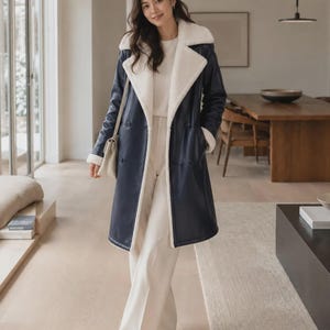 May include: A long navy blue coat with a white faux shearling lining, wide lapels, and a double-breasted button closure. The coat is worn over a white long-sleeved top and white wide-leg trousers, paired with a small cream-colored shoulder bag.