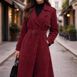 May include: A long, deep red suede trench coat featuring a double-breasted button front, a matching fabric belt with a buckle, wide lapels, and shoulder epaulets. The coat is worn over a black turtleneck and paired with black sunglasses.