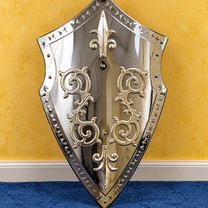 May include: A polished silver-toned metal shield featuring a pointed heater shape with a decorative border of small triangular indentations. The surface is adorned with symmetrical gold-toned scrollwork and two fleur-de-lis emblems at the top and bottom.