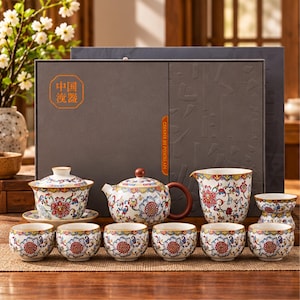 May include: A tea set featuring a teapot, a lidded bowl, a pitcher, and six small cups, all decorated with a colorful floral pattern on a white background. The set is presented in a large, dark gray rectangular box with orange text.