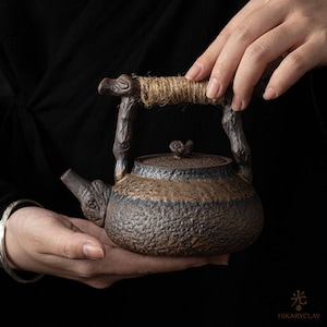 Puede incluir: Una tetera de cerámica de color marrón oscuro con una textura rugosa similar a la piedra. El asa tiene forma de rama de árbol nudosa envuelta en cuerda de yute natural. Un pequeño pomo en forma de caracol corona la tapa y el pico es texturizado.