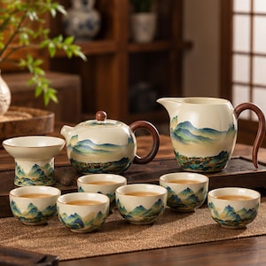 May include: A ceramic tea set featuring a teapot, a pitcher, and six small cups, all decorated with a hand-painted blue and green mountain landscape on a cream-colored base. The teapot and pitcher have dark brown wooden handles.