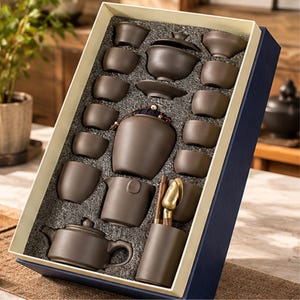 May include: A dark brown ceramic tea set arranged in a rectangular gift box with foam inserts. The set includes a teapot, a lidded gaiwan, multiple small tea cups, a tea canister, a pitcher, a utensil holder, and two gold-toned metal tools.