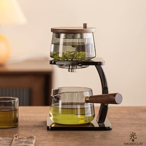 May include: A glass tea brewing set featuring a top infuser with a wooden lid and a lower glass pitcher with a wooden handle, both supported by a black stand. The set contains green tea leaves and liquid, with the text HIKARYCLAY and a symbol.