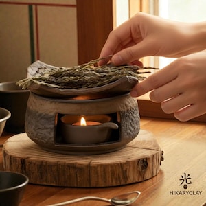 May include: A ceramic tea leaf roaster with a textured, two-tone brown and gray finish. The cylindrical base features an open chamber containing a small lit candle, while the top holds a shallow dish filled with dried green tea leaves.