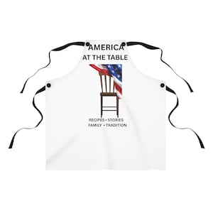 May include: A white apron with black ties featuring a graphic of a brown wooden chair draped with an American flag. The text reads AMERICA AT THE TABLE above the chair and RECIPES • STORIES FAMILY • TRADITION below the chair in black lettering.