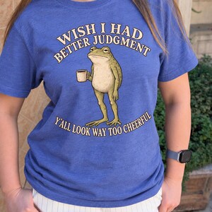 May include: A heather blue short-sleeved t-shirt featuring a graphic of a standing frog holding a small mug. The text above the frog reads "WISH I HAD BETTER JUDGMENT" and the text below reads "Y'ALL LOOK WAY TOO CHEERFUL" in a cream font.