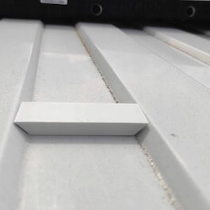 Op de afbeelding: Een wit rechthoekig onderdeel met afgeschuinde randen zit gecentreerd in een groef op een wit gegolfd oppervlak. Het oppervlak heeft parallelle ribbels en groeven, met een zwarte horizontale balk zichtbaar aan de bovenrand van het beeld.