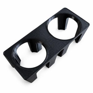 Peut inclure: Un support rectangulaire en plastique noir imprimé en 3D, doté de deux ouvertures circulaires conçues pour accueillir des objets cylindriques. La structure présente une finition texturée et repose sur quatre pieds courts et robustes.