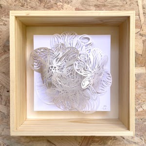 Peut inclure: Un cadre carré en bois clair de type boîte vitrine contenant une découpe en papier blanc aux motifs floraux complexes et superposés. La découpe présente des lignes délicates et des formes organiques, avec les initiales MR et 2026.