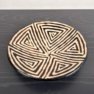 May include: A round ceramic plate featuring a high-contrast design of black and cream-colored geometric patterns. The surface is divided into triangular sections, each filled with concentric, spiraling lines that radiate from the center.