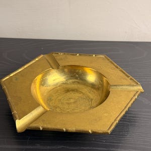 May include: A hexagonal brass ashtray with a polished gold-tone finish. The object features a central circular basin and three integrated notches along the raised, textured rim, which mimics the appearance of bamboo segments.