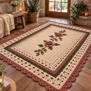 Puede incluir: Una alfombra rectangular de ganchillo con un centro de color crema y un motivo floral central de dos rosas rosadas y hojas verdes. La alfombra tiene un borde de varias capas en verde oscuro, crema y un borde festoneado en color rosa viejo.