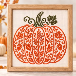 Puede incluir: Un bordado en punto de cruz cuadrado que muestra una gran calabaza naranja con intrincados patrones botánicos de hojas, bayas y bellotas. La calabaza tiene un tallo verde oscuro y una hoja, todo enmarcado en un marco cuadrado de madera clara.