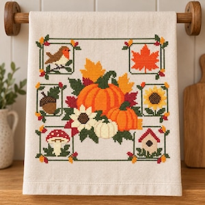 May include: A cream-colored kitchen towel featuring a cross-stitch design of autumn motifs. The central arrangement includes orange pumpkins, sunflowers, and leaves, surrounded by framed icons of a robin, maple leaf, acorn, mushroom, and birdhouse.