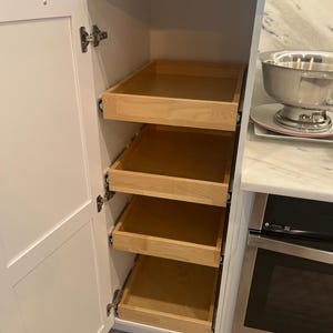 Peut inclure: Un grand placard de cuisine blanc avec une porte unique dotée d'une poignée dorée. À l'intérieur, quatre tiroirs coulissants en bois clair sont empilés verticalement, offrant un espace de rangement organisé dans le cadre du meuble.
