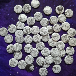May include: A collection of round, black and white pins featuring various illustrations. The pins are arranged in a star shape on a blue fabric background.