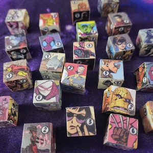 May include: A collection of cube-shaped dice featuring comic book character illustrations. Each die has six sides, each displaying a different character or number. The dice are arranged on a purple surface, creating a colorful and thematic display.