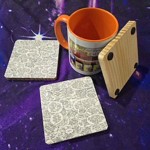 May include: A set of comic book-themed coasters with a matching orange-handled mug. The coasters feature black and white speech bubbles, and the mug has a comic strip design. A natural wood coaster is also present.