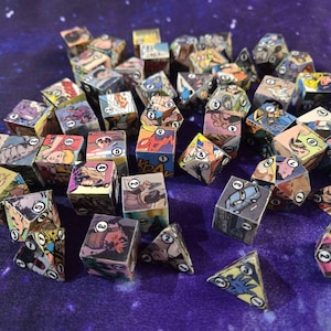 May include: A collection of comic book-themed dice. The dice are cube-shaped with comic book panels on each side, and numbered faces. The dice are scattered on a purple surface with a starry pattern.