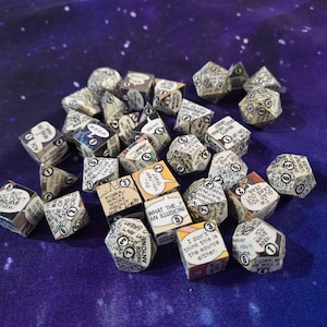 May include: A collection of multi-sided dice, each featuring comic book panels. The dice are in various shapes and sizes, with black text and numbers on a cream background. They are scattered on a purple surface.