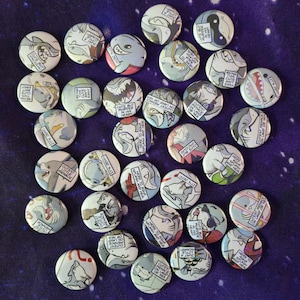 May include: A selection of circular pin badges with shark designs and text. The badges feature shark illustrations in blue, grey, red, and black, with phrases such as "Cutest Little Menace" and "Here to Aqua Splash and Heal".