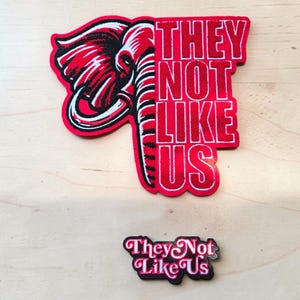 Könnte beinhalten: Ein großer rot-schwarzer Stickaufnäher mit einem Elefantenkopf und dem weißen Schriftzug "THEY NOT LIKE US" sowie ein kleinerer schwarz-weißer Emaille-Pin mit demselben Text in stilisierter Schrift, beide auf einer hellen Holzoberfläche.